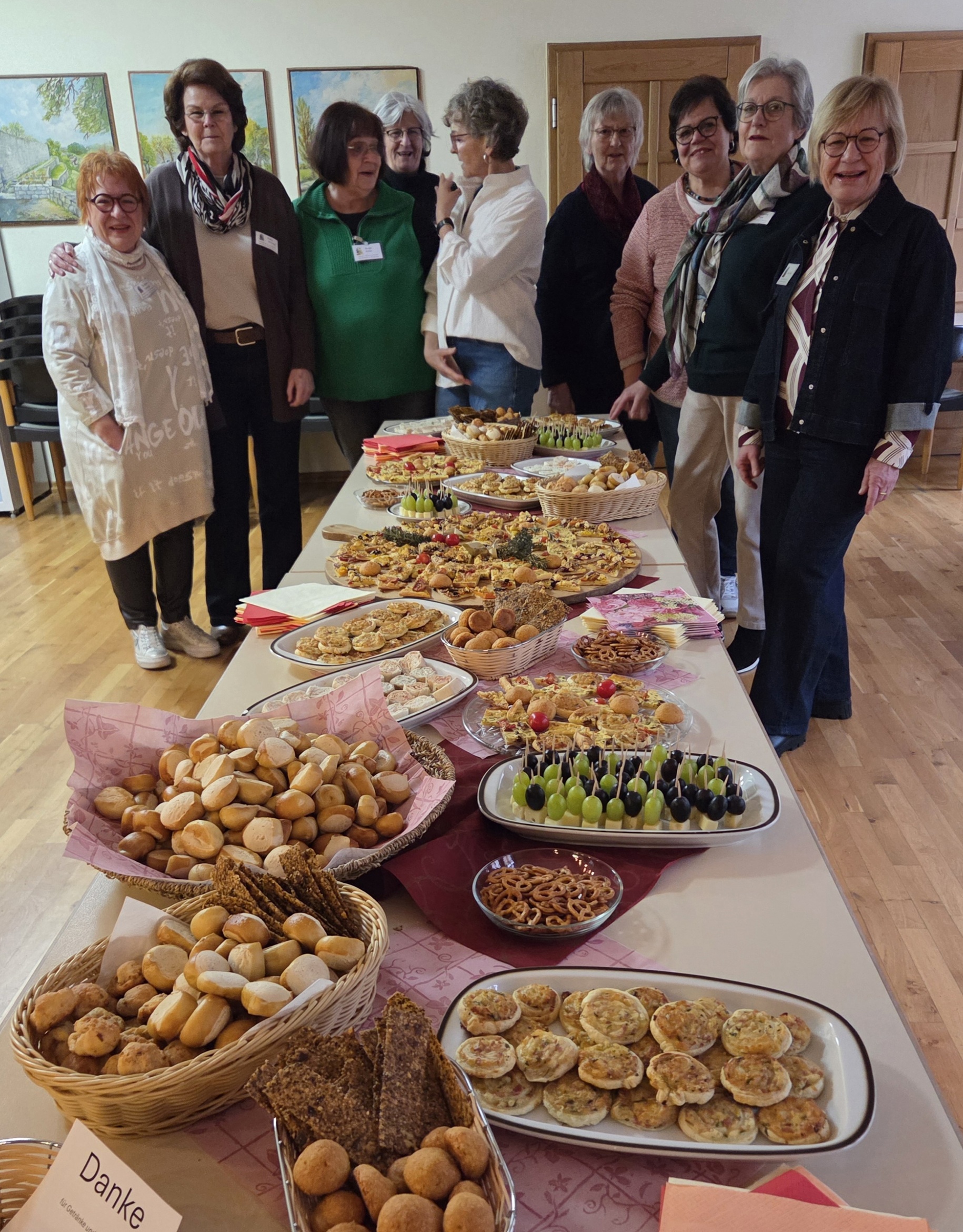 Das Bild zeigt eine Gruppe von Frauen, die hinter einem reich gedeckten Tisch stehen. Auf dem Tisch befinden sich verschiedene Speisen wie Brötchen, Kuchen, Mini-Quiches, belegte Brote und kleine Häppchen. Die Frauen lächeln und tragen Namensschilder. Im Hintergrund sind Bilder an der Wand zu sehen.