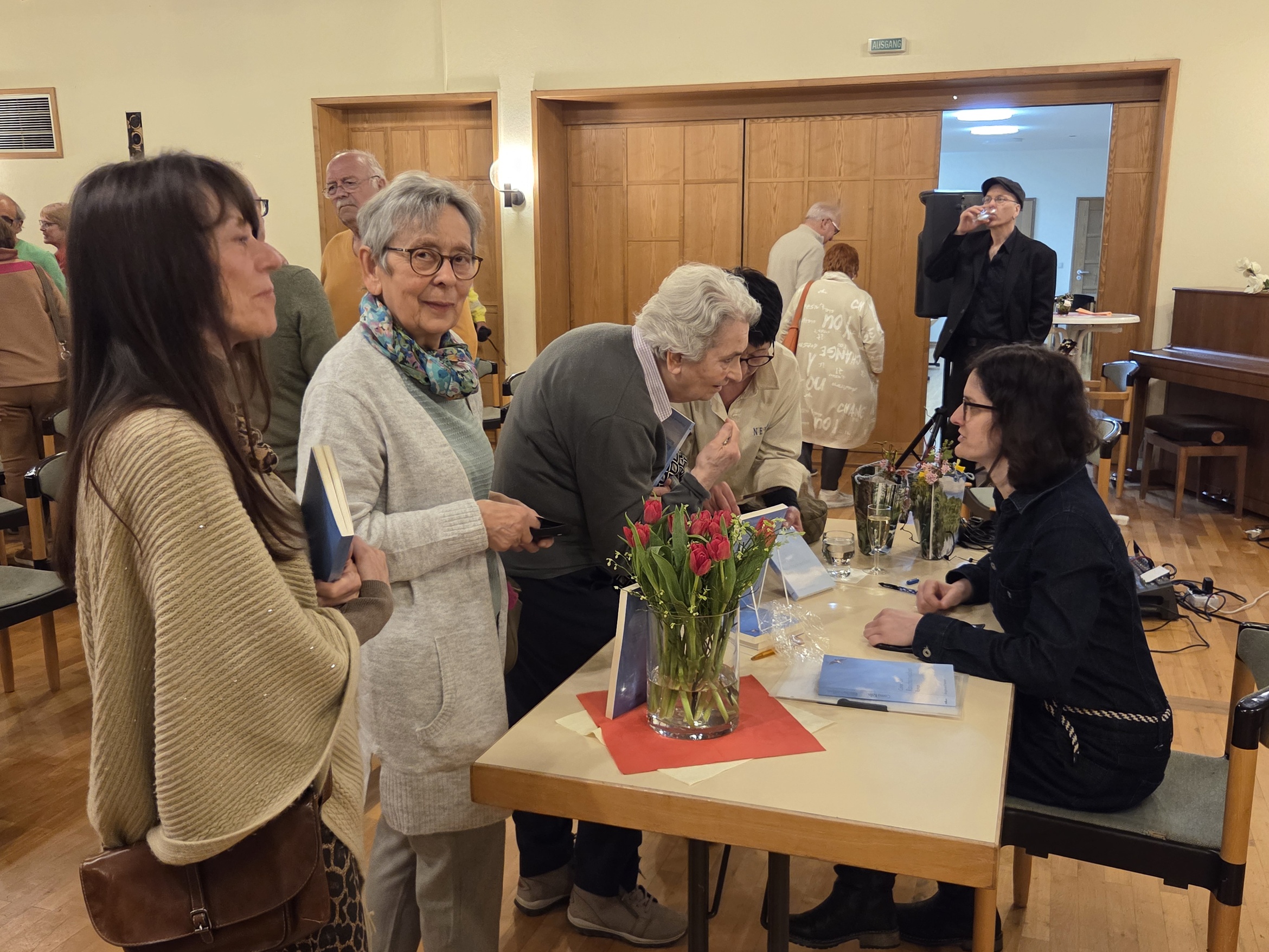 Das Bild zeigt eine Gruppe von Personen in einem hellen Raum, vermutlich bei einer Veranstaltung. Im Vordergrund stehen zwei Frauen an einem Tisch, auf dem ein Blumenstrauß und Bücher liegen. Im Hintergrund sind weitere Personen zu sehen, die sich unterhalten oder etwas betrachten. Der Raum hat Holzmöbel und eine gemütliche Atmosphäre.