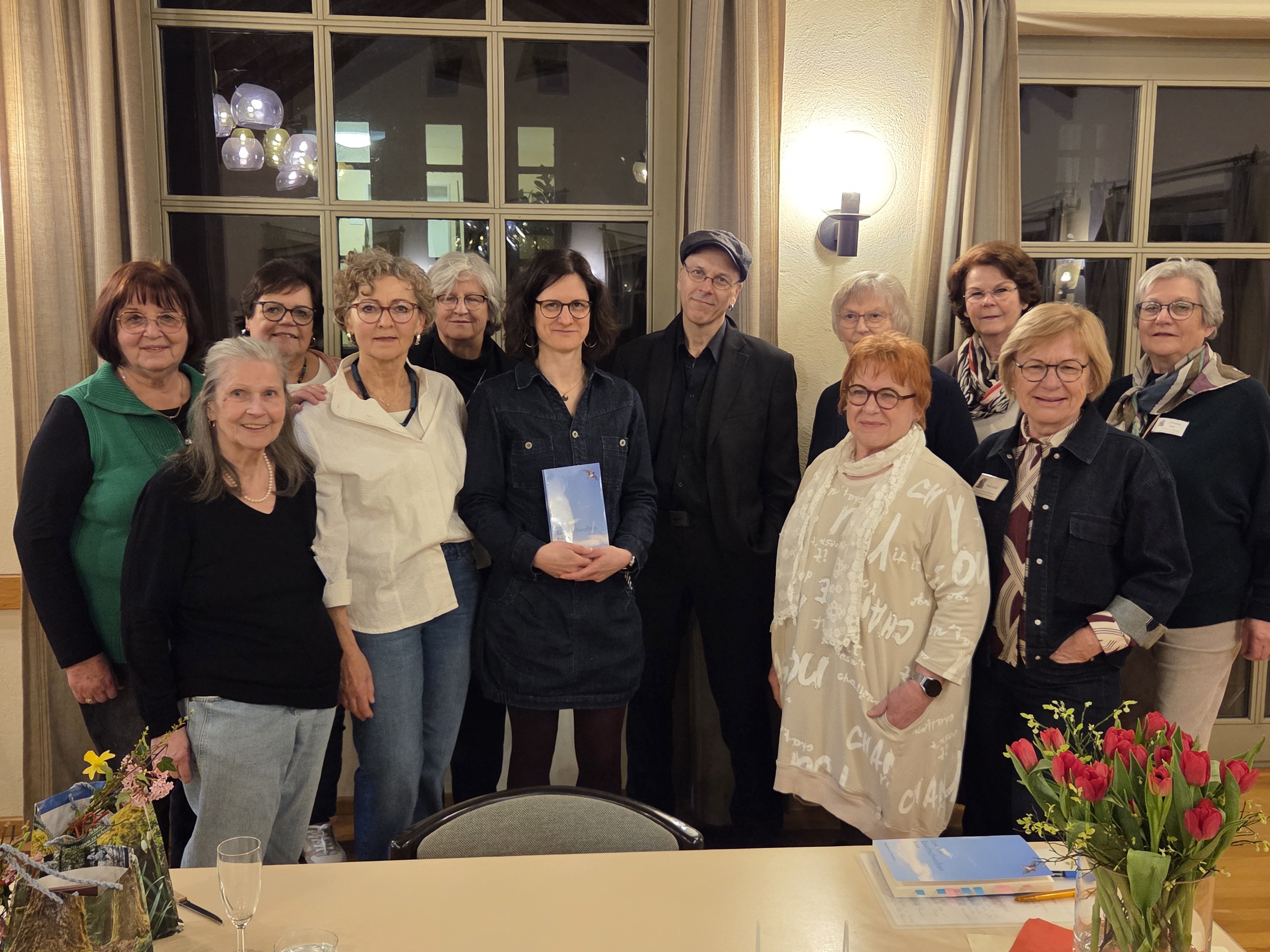 Auf dem Foto sind zwölf Personen in einem Raum zu sehen. Sie stehen in zwei Reihen vor einem Fenster und lächeln in die Kamera. Auf einem Tisch im Vordergrund stehen Blumen und einige Gegenstände.