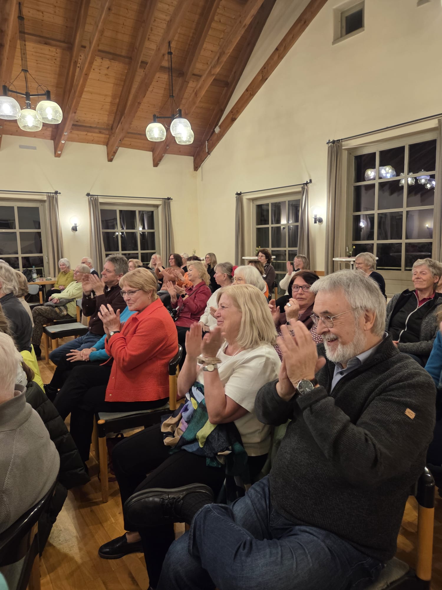 Das Bild zeigt eine Gruppe von Menschen, die in einem Raum mit Holzbalkendecke und großen Fenstern sitzen. Sie klatschen und scheinen einer Veranstaltung oder Lesung zu folgen. Die Atmosphäre wirkt warm und freundlich.