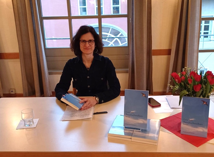 Eine Frau sitzt an einem Tisch mit mehreren Büchern, einem Glas Wasser und einem Blumenstrauß. Sie hält ein Buch in der Hand und scheint zu lesen oder zu signieren. Hinter ihr sind Fenster und Vorhänge zu sehen.
